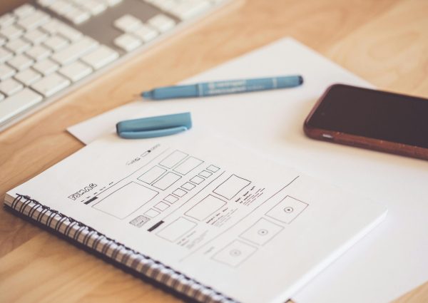 A neat workspace featuring a sketchpad with wireframes, a smartphone, and a keyboard on a wooden desk.